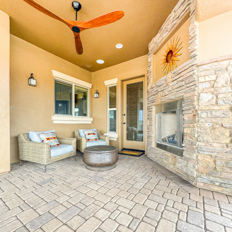 A cozy outdoor patio area at the Koehler Residence features two wicker armchairs with cushions and a small round table. A fireplace with a stacked stone finish dominates one side, while a ceiling fan with wooden blades hangs above. Well-lit with wall sconces, the space also boasts a decorative sunburst.