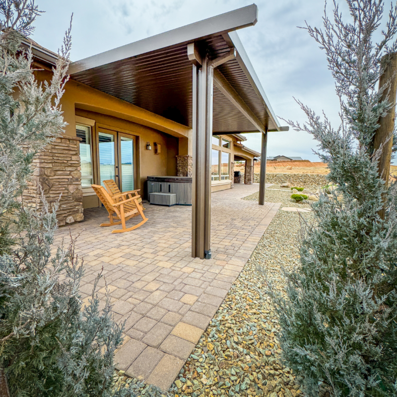 The Koehler Residence features a covered patio with a paved stone floor, showcasing a wooden rocking chair and a hot tub. The modern house boasts stone and stucco exterior walls. The patio is framed by two tall bushes, with desert landscaping visible in the background under a cloudy sky.