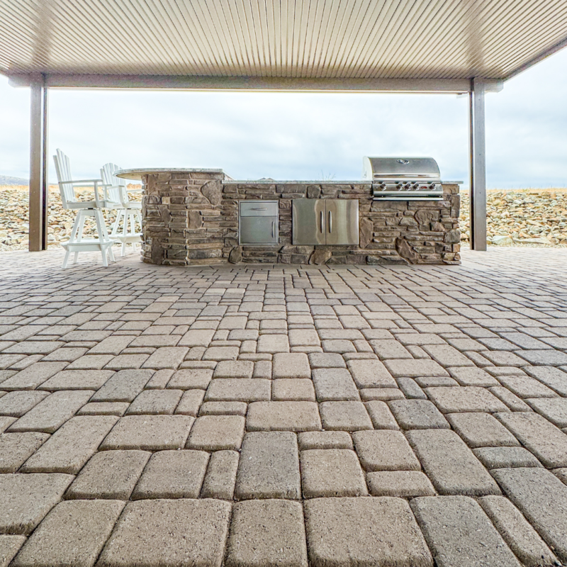 The Koehler Residence features an outdoor kitchen with a stone and stainless steel barbecue setup under a covered patio. The ground is paved with interlocking bricks, and two white chairs are placed against the stone counter. In the background, there&#039;s a low stone wall and an open sky.