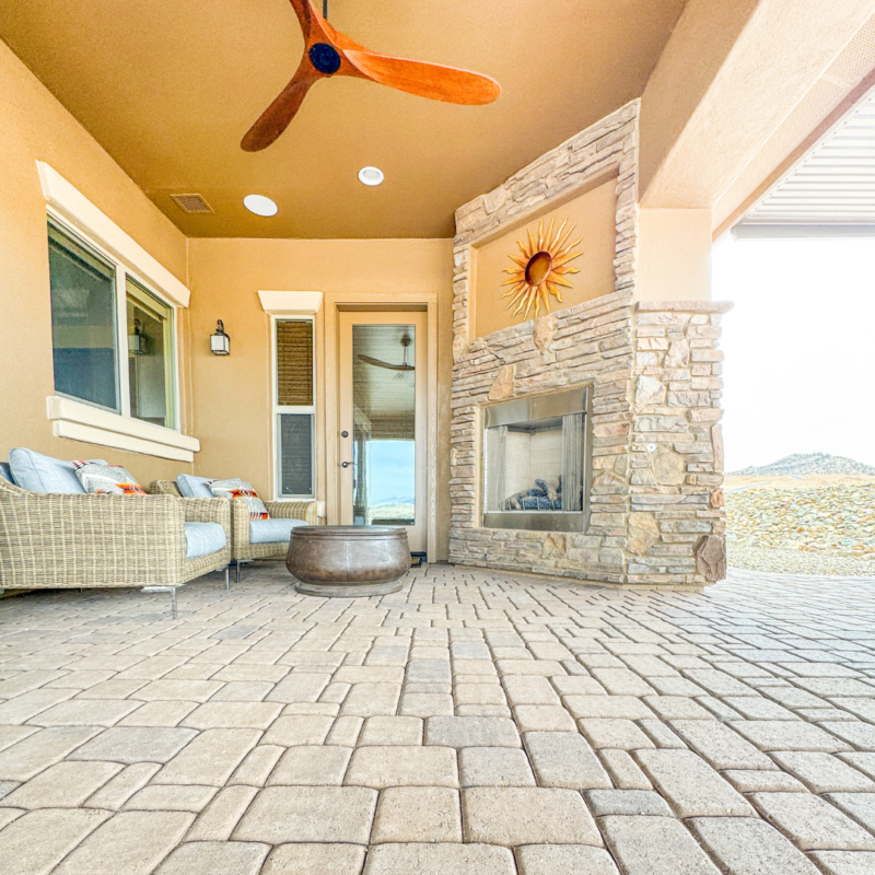 The Koehler Residence boasts a spacious, covered patio featuring wicker furniture with cushions, a stone fireplace adorned with a sunburst wall decoration, and a wooden ceiling fan. The floor is made of interlocking stone pavers, and there are mountain views in the background.