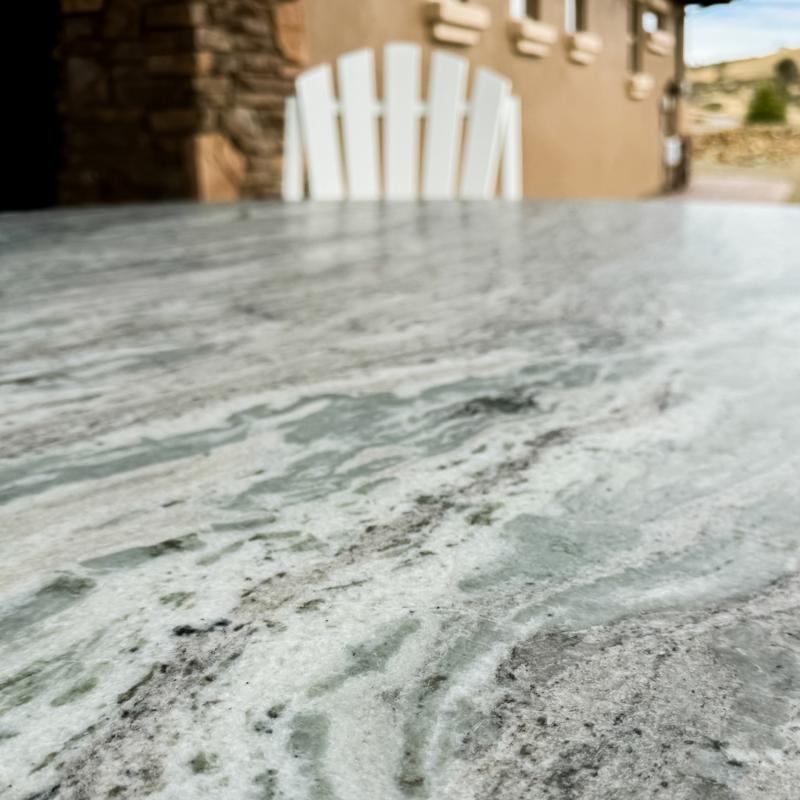 Close-up of a polished stone outdoor table with intricate swirling patterns in shades of gray and white at the Koehler Residence. In the background, a white wooden chair and part of a building with stone and stucco architecture are visible. The setting appears sunny and calm.