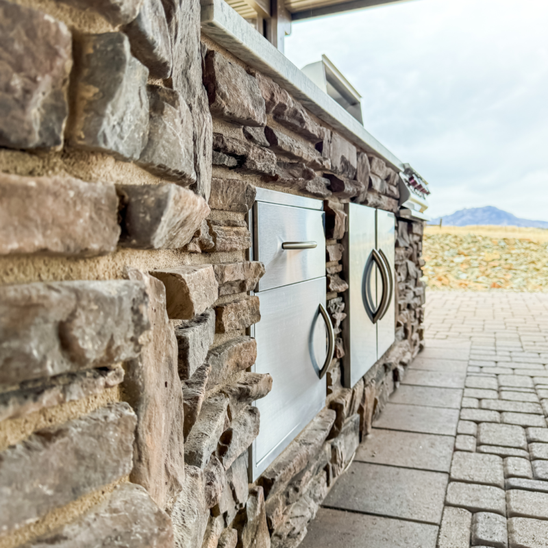 The Koehler Residence boasts an outdoor kitchen with a stone facade, featuring stainless steel drawers and a cabinet. This setup is elegantly situated on a brick patio, offering breathtaking views of rocky terrain and mountains in the background under a cloudy sky.