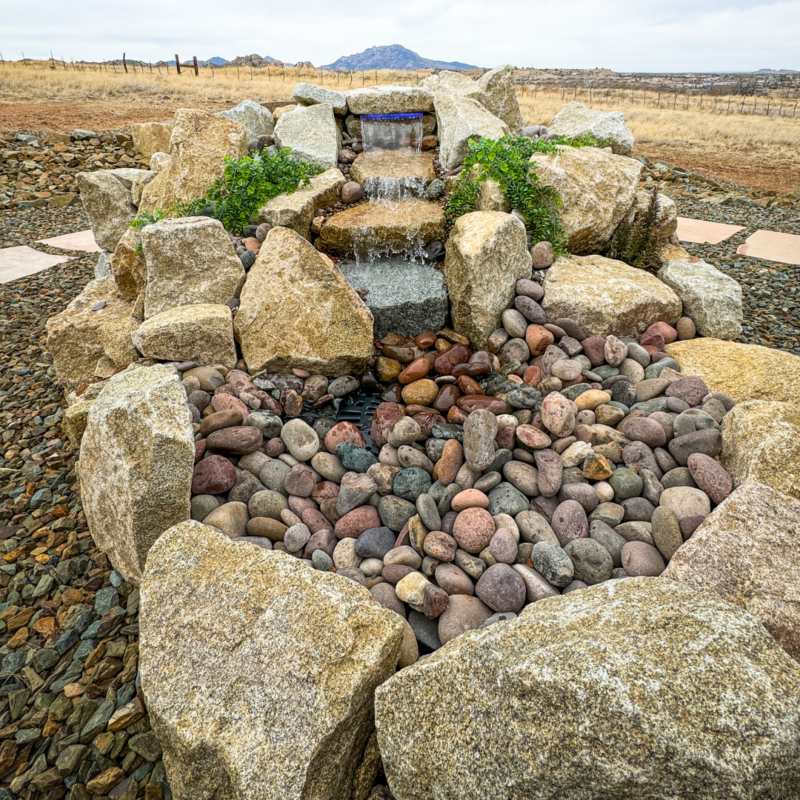 A small, serene rock waterfall at the Koehler Residence features large boulders and smooth pebbles, with a backdrop of dry, grassy plains and distant hills under a cloudy sky. The water cascades gently down the rocks into a pebble-filled basin.