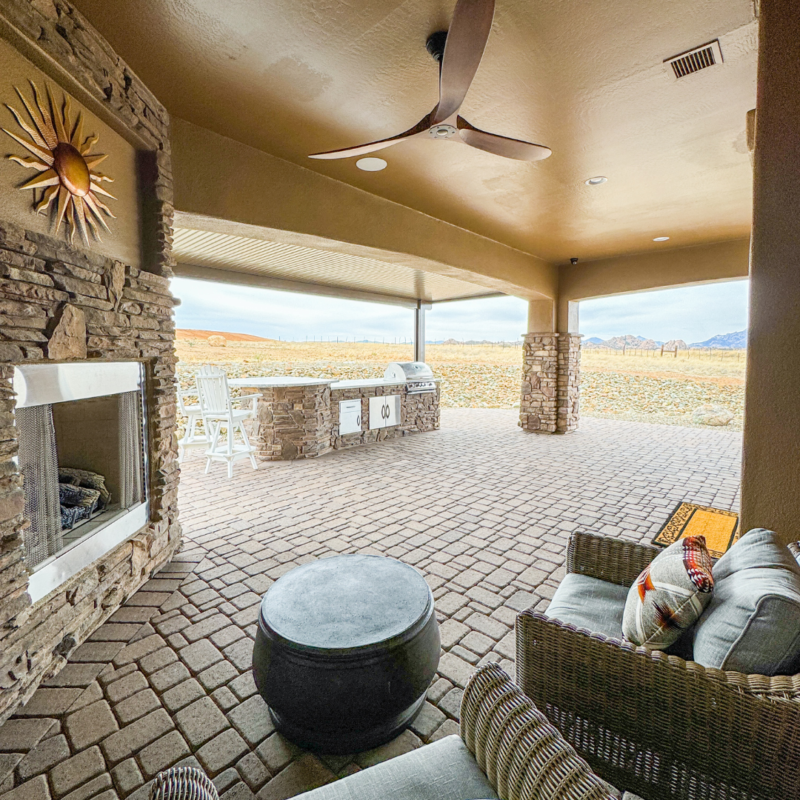 A covered outdoor patio at the Koehler residence features a cozy seating area, sun-shaped wall decor, and a fireplace on the brick floor. There&#039;s an outdoor kitchen with a grill and barstool seating in the sunny backyard, surrounded by open fields and distant mountains.