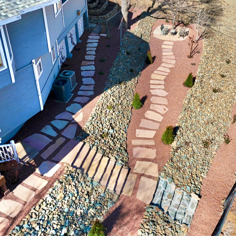 A backyard with a stone path winding through a sloped, rock-covered landscape. The path consists of irregularly shaped, flat stones, resembling an auto draft sketch in nature. There is a patio with a table and chairs on the upper right, surrounded by dry, gravelly terrain and sparse greenery.