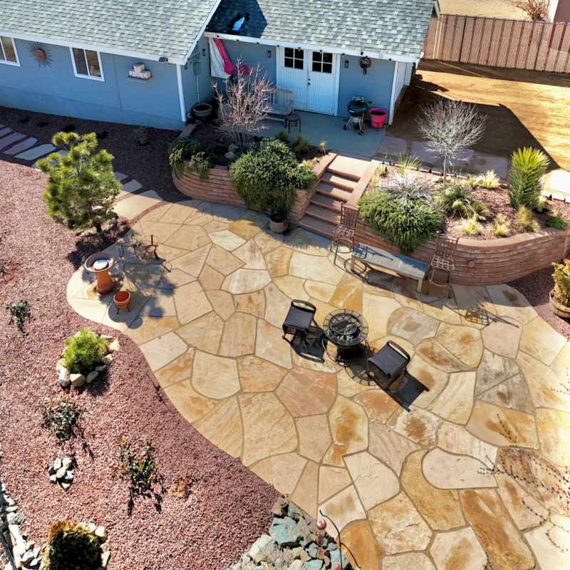 Aerial view of the Lacoss Residence backyard featuring a large stone patio with outdoor furniture arranged around a fire pit. The patio is surrounded by a well-maintained garden with various plants and shrubs. There is a blue house with a gray roof in the background.