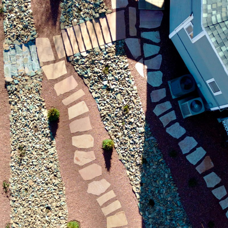 Aerial view of the Lacoss Residence corner with a stone pathway on a rocky landscape. The path, made of irregular stone slabs, curves through the yard. Two air conditioning units are adjacent to the house with a shadow cast over part of the area.