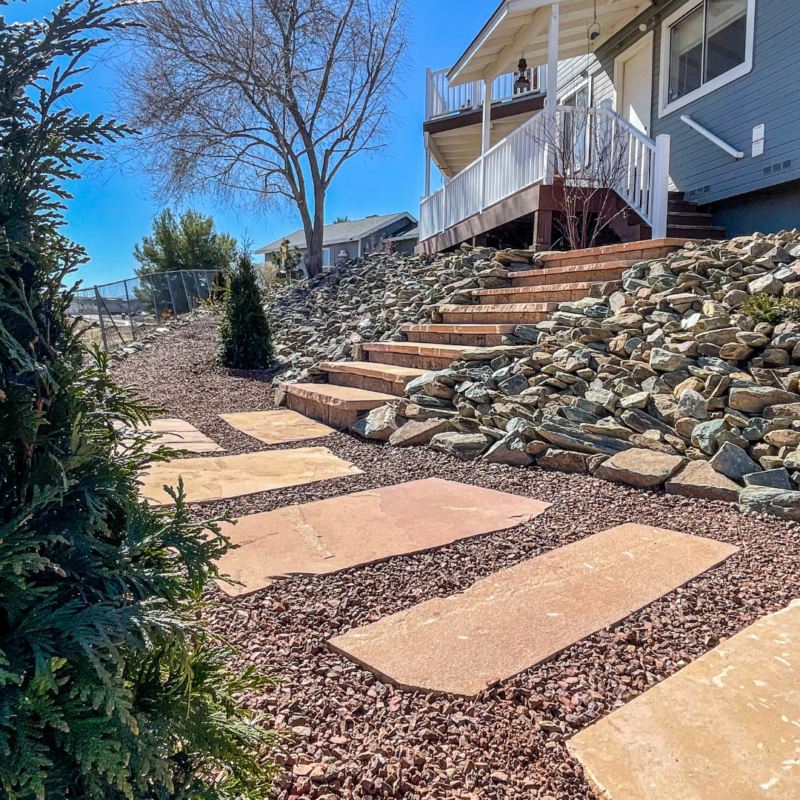 A pathway made of large, flat stones leads to steps ascending to the porch of the Lacoss Residence. The yard features landscaped rock coverage with evergreen shrubs. The house boasts a white railing around the porch and a leafless tree nearby, all under a clear blue sky.