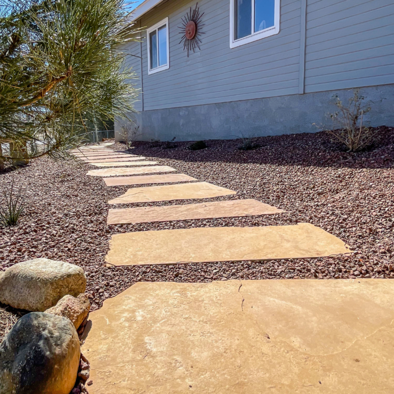 A stone pathway leads to the side of the Lacoss residence. The pathway consists of rectangular stones set against reddish-brown gravel. A plant with long green leaves is in the foreground, and a decorative sun hangs on the house&#039;s exterior wall.