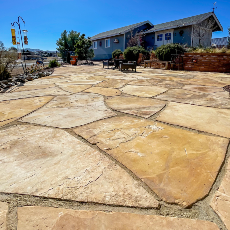 The Lacoss Residence boasts a spacious backyard patio with large, irregularly shaped stone tiles. Two bird feeders on poles are visible on the left, while a house with a gray exterior and outdoor seating area is in the background. Green vegetation lines the patio edges, and the sky is clear and blue.