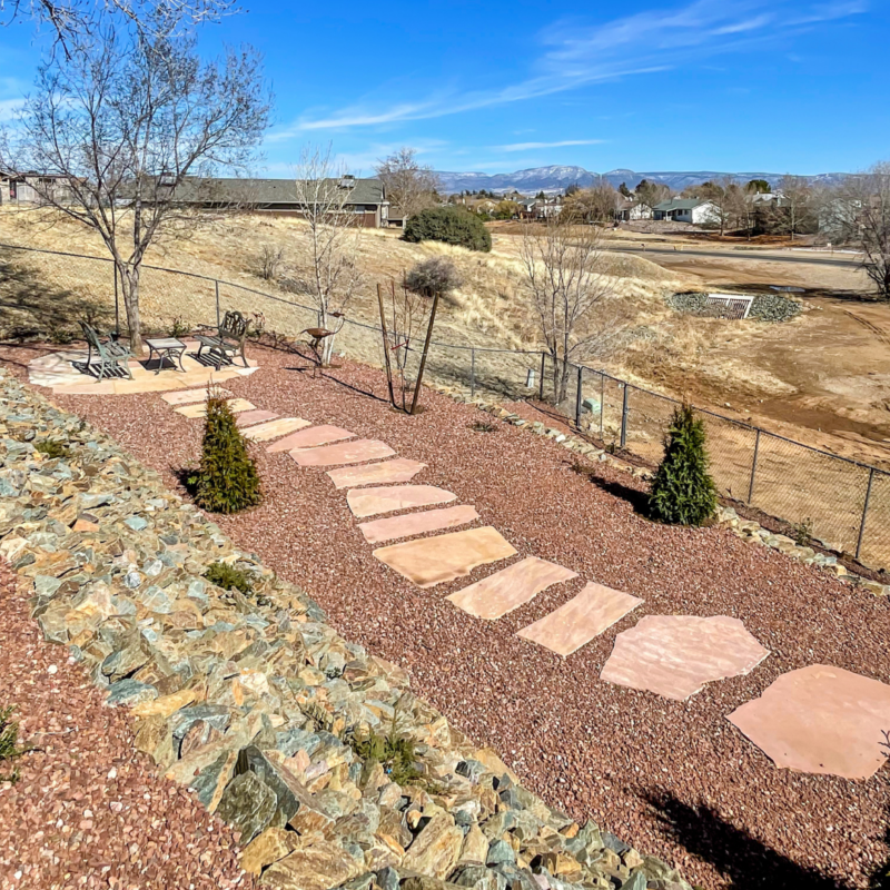 A backyard at the Lacoss Residence features a stone path leading to a seating area complete with chairs and a table. The landscape is composed of reddish gravel, a few trees, and shrubs. A fence separates the yard from an open field, with houses and mountains visible in the background.