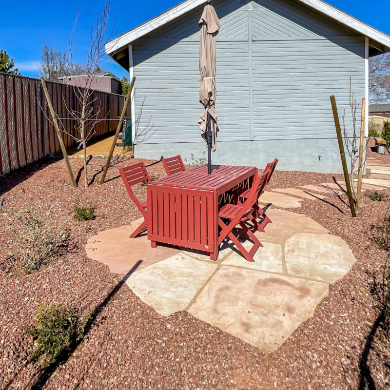 The Lacoss Residence boasts a backyard patio featuring a red wooden table and four matching chairs centered under a closed beige umbrella. The patio is surrounded by small rocks and young, leafless trees, with the light blue house in the background against a clear blue sky.