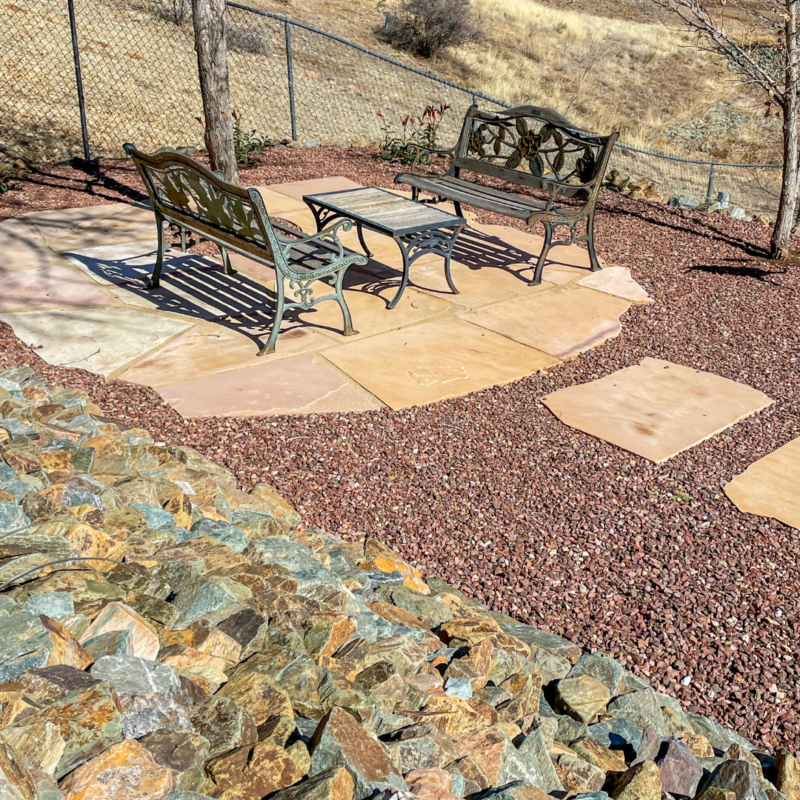 The serene outdoor seating area at the Lacoss Residence features two ornate metal benches facing each other with a small matching table in between. The stone-tiled area is surrounded by red gravel, set against a backdrop of a chain-link fence and dry, grassy terrain.