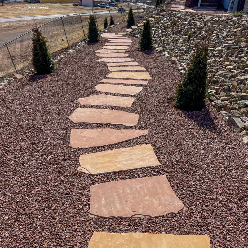 A stone path made of irregular, beige slabs set in reddish-brown gravel winds its way through the Lacoss Residence yard. Small evergreen trees line one side of the path, and a rocky embankment is on the other side. A chain-link fence runs along the far edge.