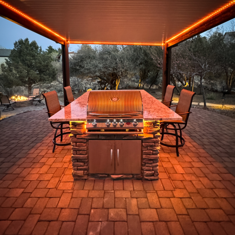 Outdoor kitchen with a built-in stone barbecue grill under a pavilion, surrounded by six patio chairs. The area is illuminated with warm, orange LED lights, creating a cozy atmosphere for the residence, while a firepit and trees are visible in the background.