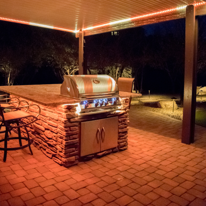 The Mcafee Residence boasts a well-lit outdoor patio at night, featuring a built-in stone barbecue grill under a pergola. The orange ambient lighting casts a warm glow over the brick floor and surrounding chairs, creating a cozy atmosphere for evening gatherings. Trees are visible in the background.