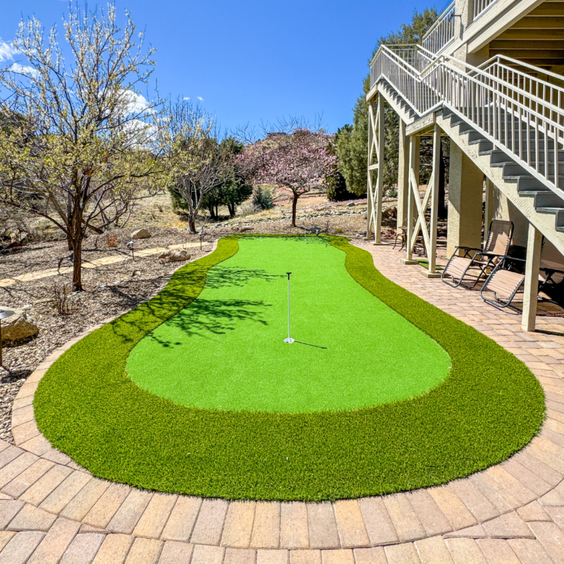 The McAfee residence features a backyard putting green surrounded by a stone-paved patio. This well-maintained green has a small flagstick at its center. Deck chairs and a stairway to an elevated deck are visible on the right, and trees with sparse foliage border the area.