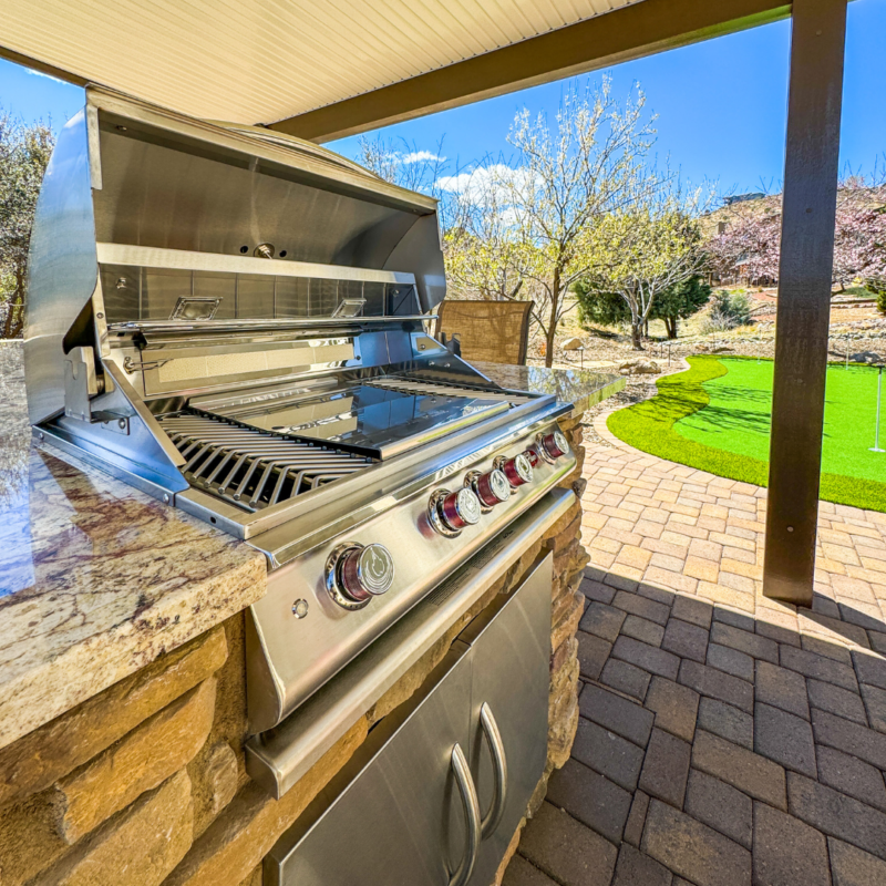 A shiny stainless steel outdoor grill with a granite countertop sits under a pergola on the stone-paved patio of the McAfee residence. In the background, well-maintained trees, a lush green lawn, and a small putting green area enhance the serene setting under a clear blue sky.