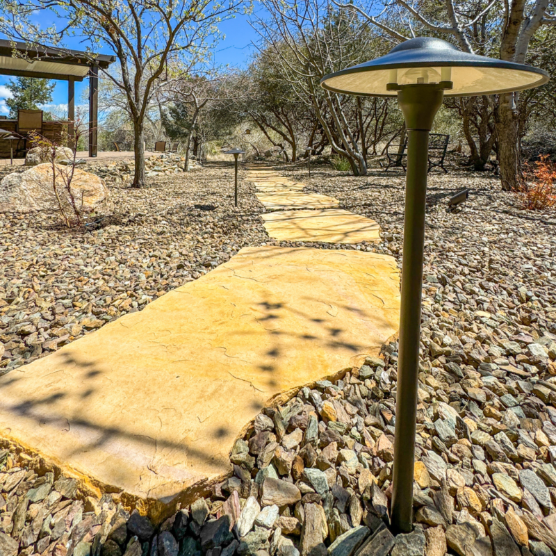 A stone pathway winds through a rocky garden, bordered by trees and shrubs. A black outdoor lamp post stands in the foreground, casting a shadow on the path. A covered patio structure is visible in the background under a blue sky, enhancing the peaceful ambiance of this picturesque residence.