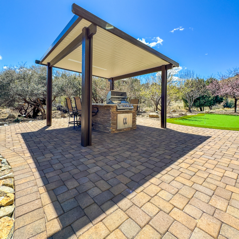 The Mcafee Residence boasts a shaded outdoor kitchen featuring a pergola-covered grilling area with stainless steel appliances, surrounded by a stone countertop. It is set on a stone-paved patio with bar stools on one side. The area is surrounded by greenery and trees under a clear blue sky.