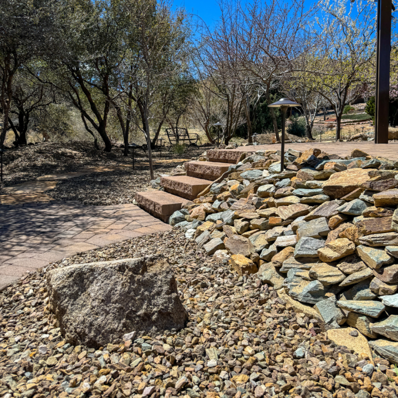 An outdoor scene at the Mcafee residence features a stone pathway leading to steps made of large bricks, surrounded by various trees and shrubs. To the right, there&#039;s a sloped area covered in assorted rocks with a small landscape light. The background is framed by clear blue skies and sunlight.