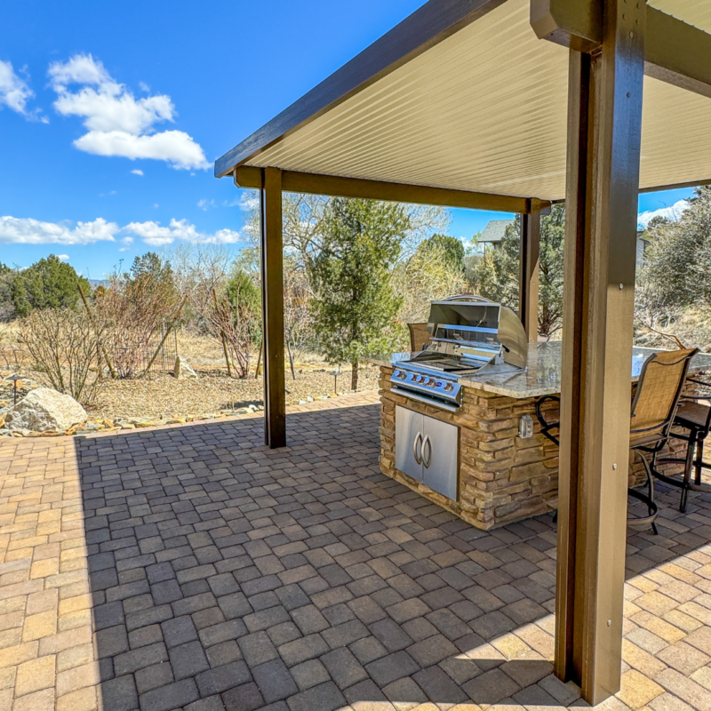 A backyard area at the McAfee residence boasts a covered patio with a built-in stone grill and countertop. The ground is paved with brick, and there are chairs around the counter. The patio overlooks a natural, rustic landscape with trees and shrubs under a bright blue sky.