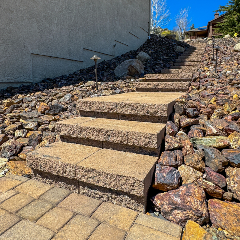 A set of outdoor stone steps leads up a sloped terrain with brown and red decorative rocks on either side. A stucco wall is on the left, and a few pathway lights are along the steps. In the background, the McAfee residence and trees are visible under a clear blue sky.