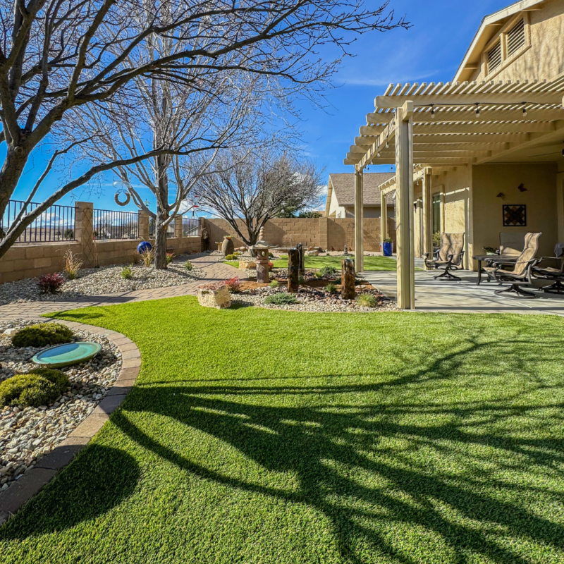 The Radil Residence boasts a well-maintained backyard with green artificial grass, small garden areas with rocks and plants, shaded seating under a pergola, and a tire swing hanging from a tree. A stone pathway runs through the garden, and wooden poles are placed decoratively in the center.