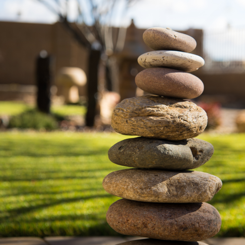 A stack of smooth stones balanced on top of each other in a garden at a serene residence. The stones vary in size and color, creating a natural and calming arrangement. The background is slightly blurred, showing green grass, a tree, and parts of a fence and building.
