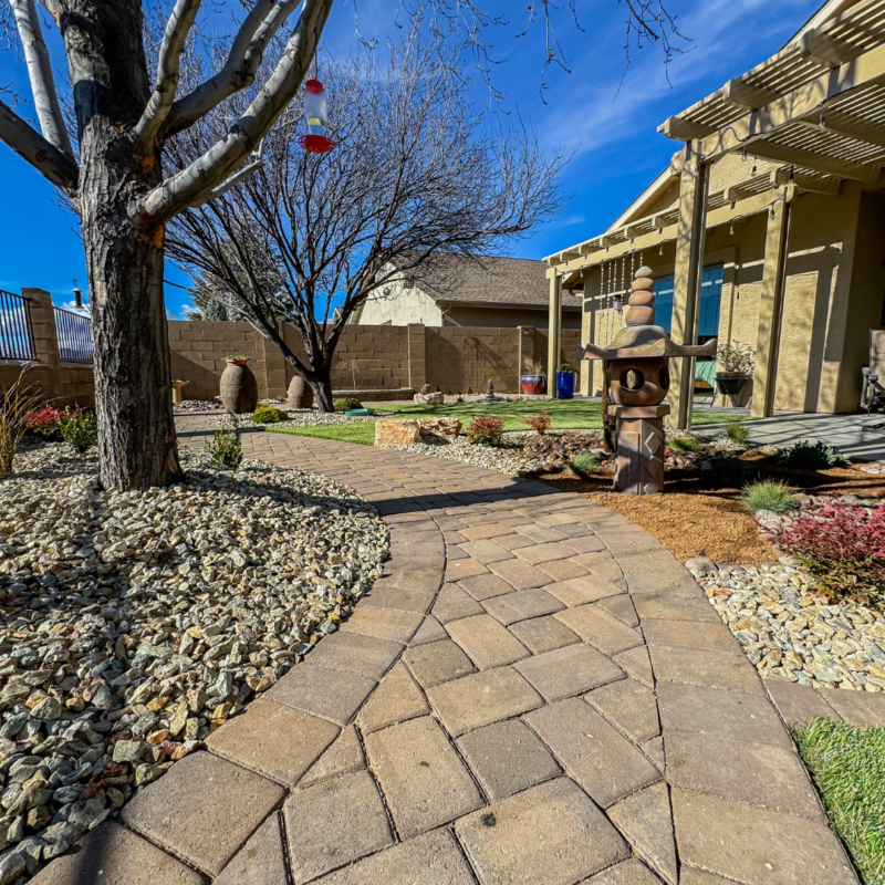 The Radil Residence showcases a landscaped backyard featuring a curved stone pathway, a tree with bare branches, a bird feeder, and decorative garden elements. The patio area has a trellis for shade, and the house is partially visible under a clear blue sky.