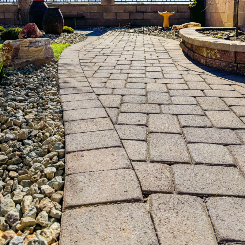 A sunlit stone pathway with a pattern of rectangular pavers winds through a garden at the Radil residence. On the left, there are small rocks and a few decorative pots. The pathway curves to the right, bordered by a short stone wall and more greenery.