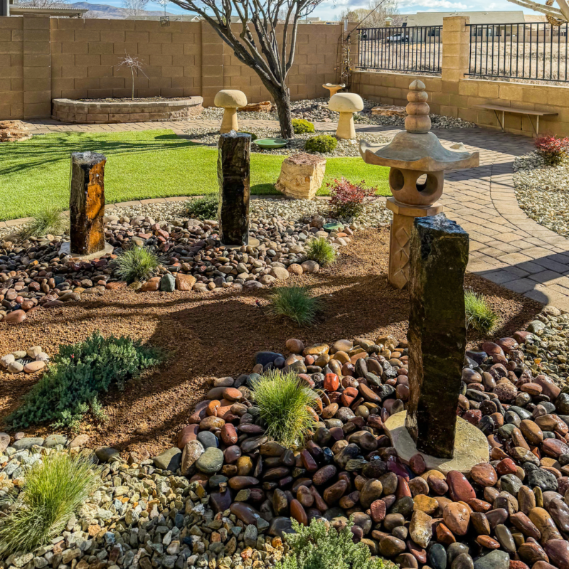 The Radil Residence boasts a serene garden with stone sculptures, varied plants, and a paved walkway surrounded by gravel. Vertical rock formations and a water feature enhance the dry, well-cared-for landscape under sunny skies.