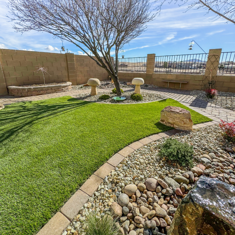 The Radil Residence boasts a landscaped backyard featuring a leafless tree surrounded by rocks and pebbles. Green artificial grass bordered by stones, two mushroom-shaped stone sculptures, a wooden bench, and a brick wall fence create a serene atmosphere. Bird feeders hang from poles in the background.