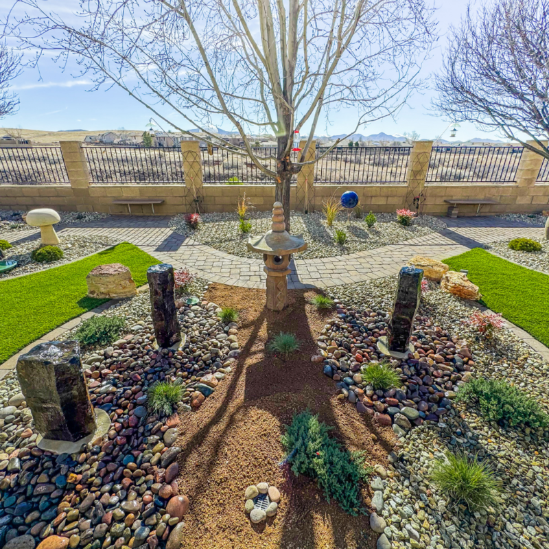 The Radil Residence boasts a serene backyard garden featuring a tree at the center, surrounded by stones and small plants. Japanese-inspired stone lanterns and decor elements are placed around the garden. The area includes patches of green grass and a stone pathway, framed by a stone fence.