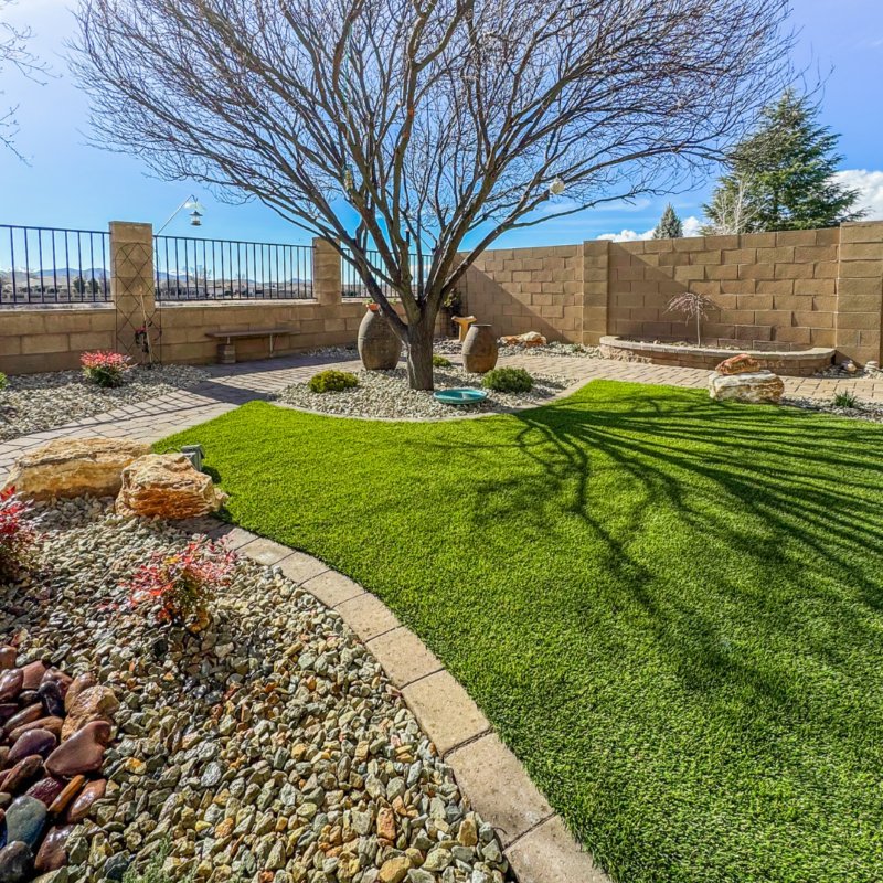 The Radil Residence boasts a well-maintained yard featuring a tree at the center with a stone mulch bed around it. The lush, green artificial grass is bordered by light-colored stones in the foreground. Enclosed by a brick wall, the yard is complete with garden decor and a cozy bench along the sides.