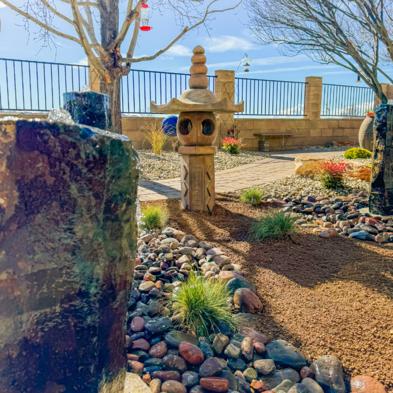 A serene Japanese garden at the Radil Residence features a stone lantern surrounded by a mix of pebbles and small plants. Two large stones frame the pathway leading to the lantern. Leafless trees and a clear blue sky complete the peaceful setting.