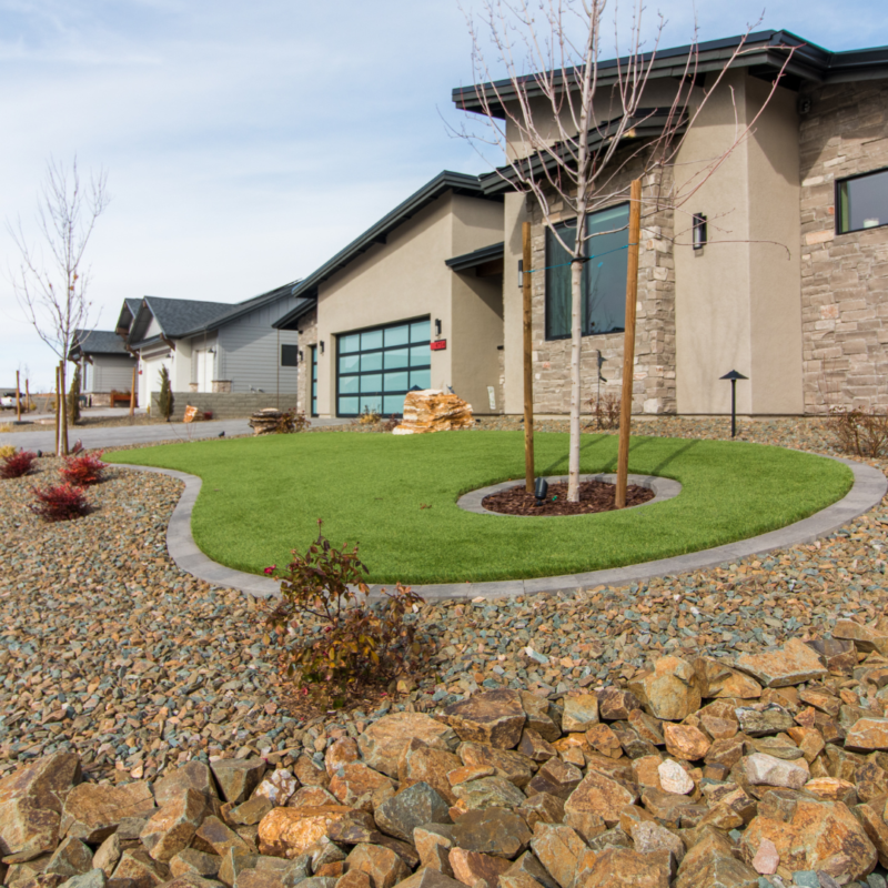 Front yard of the Schow Residence, a modern house with beige and light gray exterior. The yard features a well-maintained green lawn, young trees, and surrounding rocky landscaping. Two other houses are visible in the background, lined up along a suburban street.