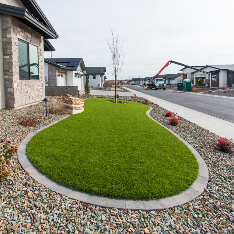 A neatly landscaped front yard at the Schow Residence features a spacious, well-manicured green lawn and a young tree in the center, surrounded by gravel and stone paths. Modern houses and a street lined with construction vehicles are visible in the background.