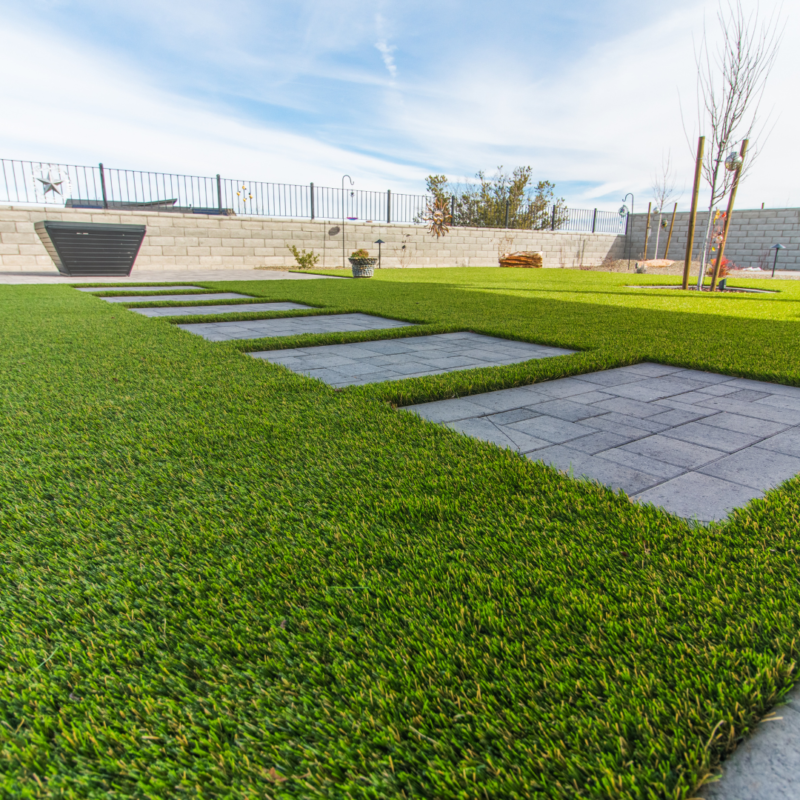 The Schow Residence boasts a backyard with a well-maintained green lawn and a paved stone pathway leading through it. A charming stone wall encloses the area, dotted with trees and plants near the perimeter. The sky above is clear with just a few clouds.