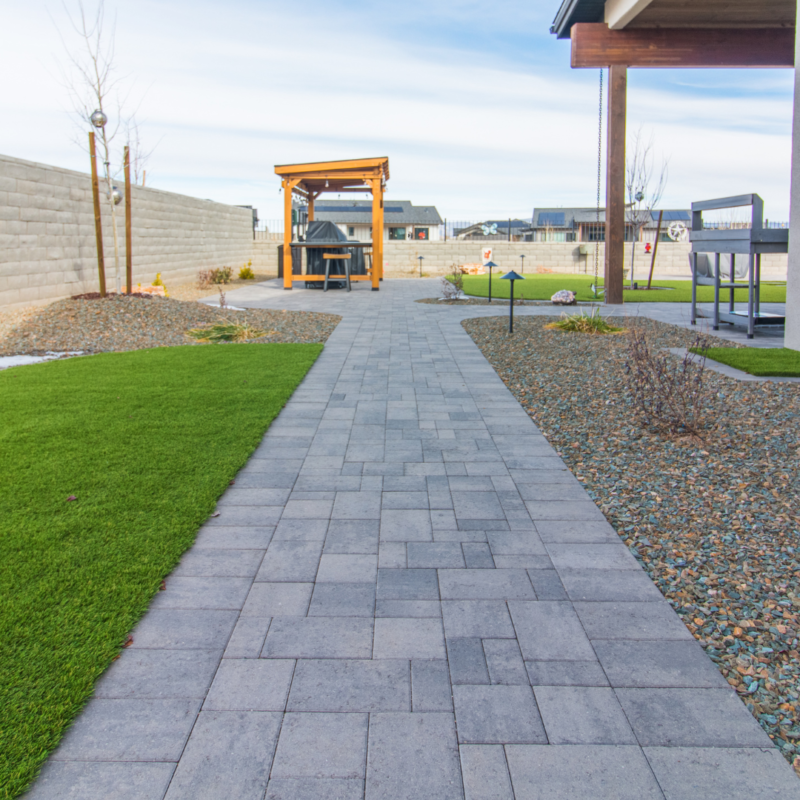 A backyard at the Schow Residence features a stone tile pathway leading to a covered wooden pergola with a grill and seating area. The pathway is flanked by green grass patches and gravel landscaping. In the background, a concrete block fence encloses this tranquil space.