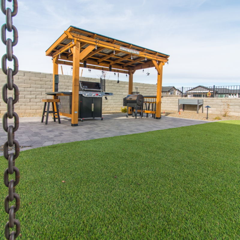 The Schow Residence features a wooden pergola with a metal roof, covering an outdoor kitchen equipped with a grill and smoker on a paved patio. A brick wall sets the background while neatly trimmed green artificial grass adorns the foreground.