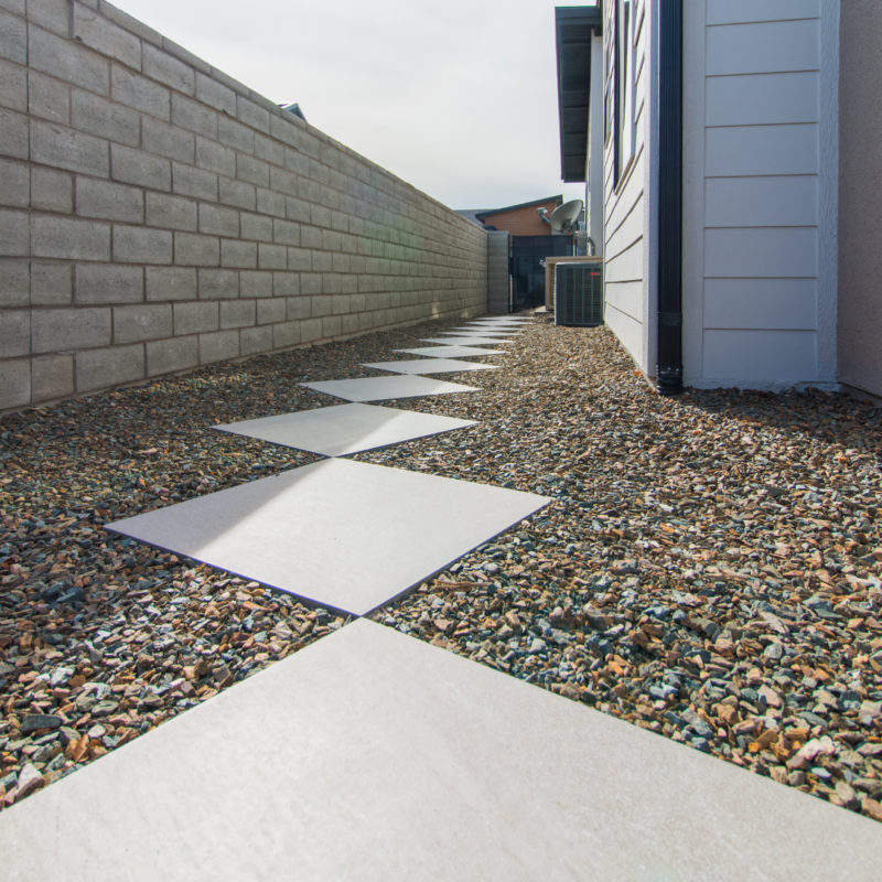 A narrow pathway along the side of the Schow Residence features diamond-shaped tiles set in gravel. The path is bordered by a beige brick wall on one side and the house wall with white siding on the other. An air conditioning unit is visible further down the path.