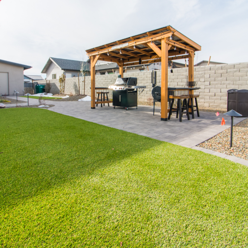 The Schow Residence boasts a backyard featuring a well-maintained lawn, a stone patio, and a wooden pergola equipped with a grill and seating area. A few houses and a stone wall are visible in the background, with clear skies overhead.