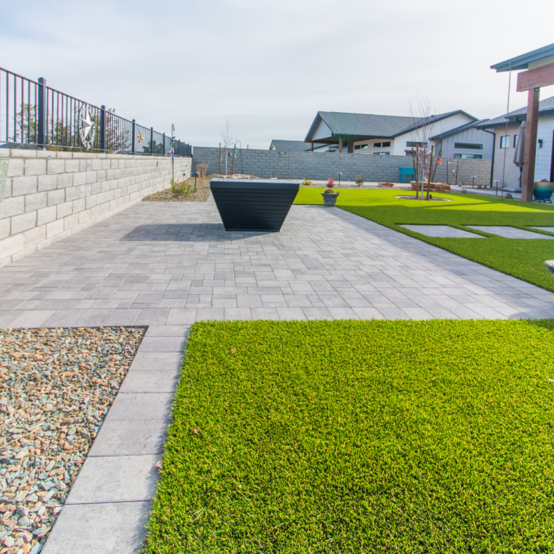 The Schow Residence features a modern, landscaped backyard with a large paved patio area, synthetic grass, and a central black planter. The space is surrounded by a mixture of wooden and stone fences, with neighboring houses visible in the background under a bright sky.