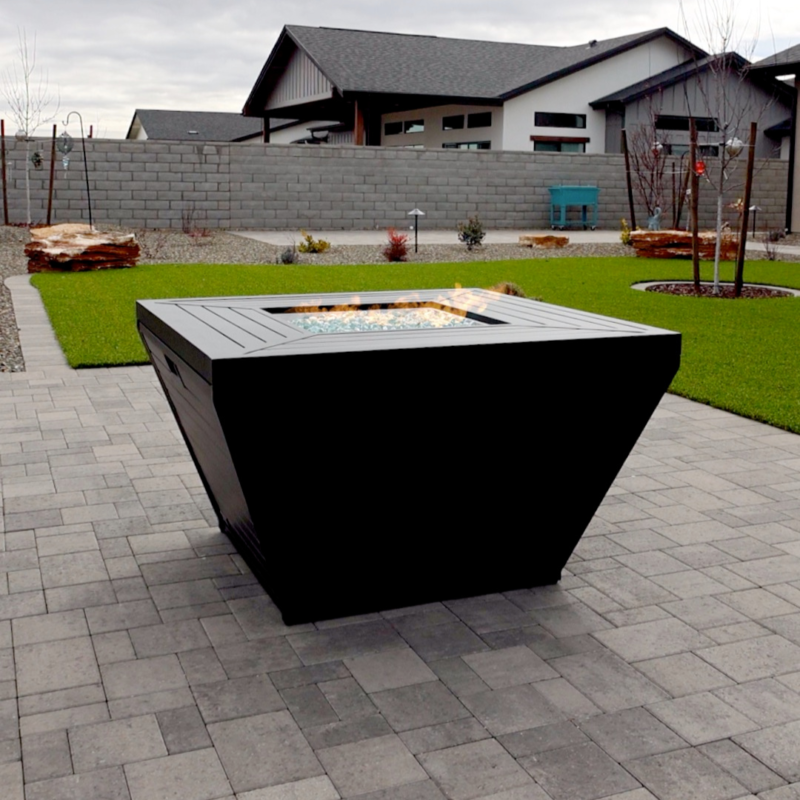 A backyard at the Schow Residence features a modern fire pit in the foreground, set on a paved patio. Flames rise dramatically from its center. The background showcases a neatly manicured lawn, a stone fence, and a few modern houses under an overcast sky.