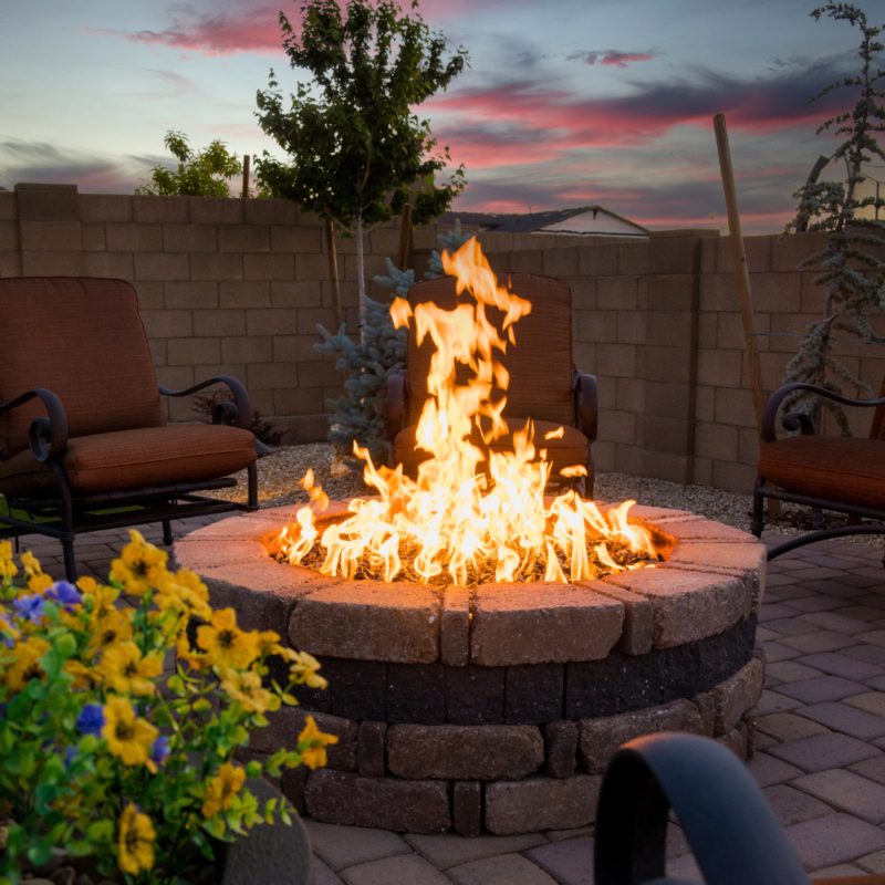 A backyard patio at the Kann Residence features a stone fire pit at dusk. The fire pit is surrounded by four cushioned chairs. Yellow flowers in a planter are in the foreground, while the sky displays a colorful sunset with purple, pink, and orange hues.