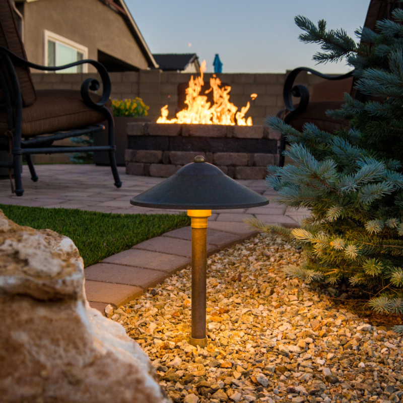 A landscaped backyard at dusk at the Kann Residence features a lit pathway light in the foreground and a cozy fire pit with flames in the background, surrounded by seating. The scene includes a mix of greenery, rocks, and pavers, creating a tranquil outdoor setting.