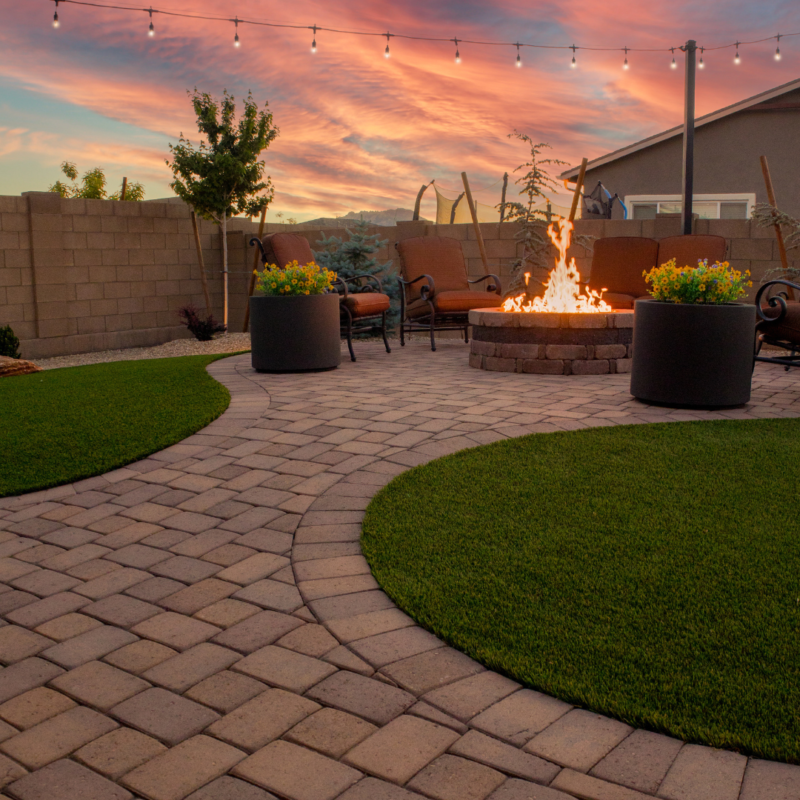 The Kann Residence&#039;s backyard patio at sunset features a circular fire pit with a glowing flame, surrounded by cozy chairs. String lights hang above, illuminating the area. Potted plants add greenery, and the sky is painted with vibrant hues of orange, pink, and purple.