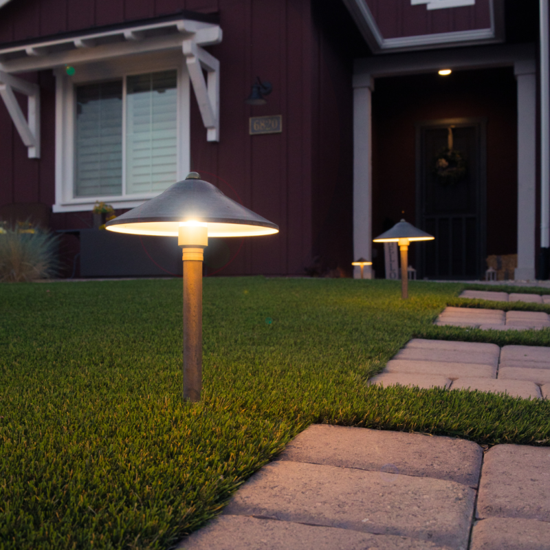 A view of the Kann Residence&#039;s front yard at dusk, featuring a pathway of stone pavers bordered by well-manicured grass. Modern, mushroom-shaped outdoor lights illuminate the path. The house has a red exterior with white trim and a dark front door.