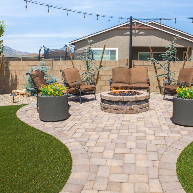 The Kann Residence backyard patio boasts patterned paving stones and a circular fire pit at its heart. Four cushioned chairs encircle the pit, perfect for cozy gatherings. Potted plants and strung lights add charm, while a grassy area and fence provide a serene backdrop.